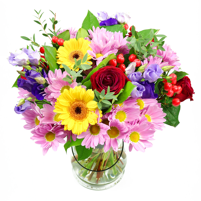 In this photograph, a generously sized, mixed-colour bouquet rises from a transparent glass vase set against a bright white backdrop, allowing every petal and leaf to stand out distinctly. The composition is anchored by sumptuous deep red roses clustered near the centre, their tightly layered, velvety petals giving the design a sense of depth and timeless romance. Close by, sunny yellow gerbera daisies with dark brown-orange centres introduce a flash of exuberant light, their wide, open faces creating a friendly, approachable mood. Surrounding these star blooms are numerous soft pink daisy-like chrysanthemums, each with a yellow eye and a halo of slender petals, building a gentle cushion of colour that makes the arrangement feel lush and generous. Interspersed between the pinks and yellows, stems of purple and pale lavender lisianthus stretch upward, their bell-shaped blooms and slightly ruffled edges providing a cool counterpoint and lending an air of contemporary elegance often favoured by clients in converted warehouse apartments around King's Cross and Coal Drops Yard. Threaded discreetly among the flowers, rounded red hypericum berries cluster along fine stems, offering pops of smooth, glossy texture and a slightly playful note. The bouquet is supported by a mix of greenery: broad, dark green leaves that give structure, and lighter, more delicate sprigs that soften the outline and suggest fresh growth. Inside the clear vase, you can see the carefully trimmed stems criss-crossing in clear water, a detail that reflects the practiced hand of an experienced florist. The overall effect is abundant yet balanced, perfect for a celebratory delivery to a new office near St Pancras, a thank-you gesture along Pentonville Road, or simply to bring a burst of colour into a Kings Cross home.